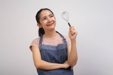 A smiling woman in a blue apron holds a whisk, looking thoughtfully upward, suggesting creativity in cooking or baking.