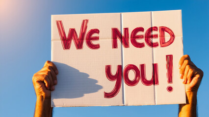 Hands Holding "We NEED You!" Sign, A powerful image of dark-skinned hands firmly holding a handmade sign displaying the bold text "We NEED you!" against a clear blue sky.