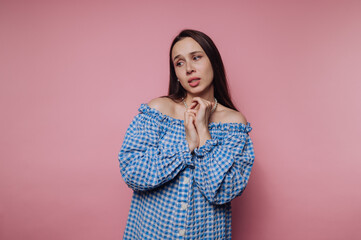 Lady in blue checkered dress poses in front of pink background with a thoughtful expression,...