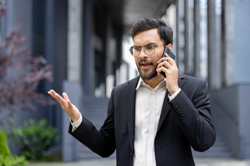 A man in business attire stands outside using a smartphone, expressing frustration during a phone...