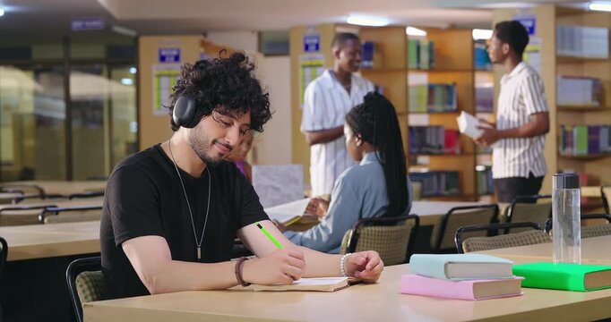Indian asian young adult gen z male man calm peace mind student sitting desk table listening music hold pencil write exam notes smart boy do self study read book prepare topics indoor library busy day