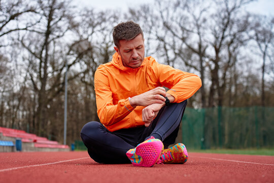 Man in sportswear sitting on red running track and checking his smartwatch. Concept of using digital gadgets in fitness training