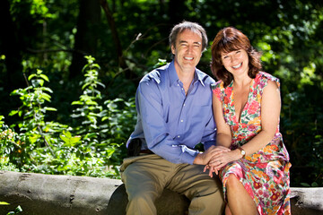 A mature couple sitting closely together holding hands in a sunlit woodland park, both smiling brightly at the camera. 