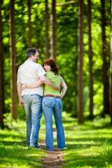 Fototapeta premium A mature man and woman on a forest path with their arms around each other. The woman looks back and smiles at the camera. From a series of images.