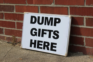 A sign that says dump gifts here leaning against a brick wall on a concrete surface outdoors