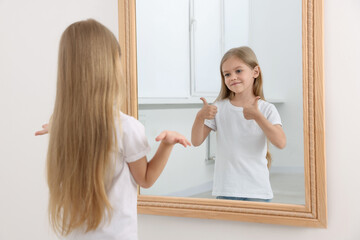 Confused little girl in front of mirror where her reflection showing thumbs up indoors. Mental...