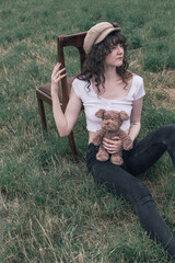 Young woman with curly brown hair wearing a white crop top, black jeans, and a beige woven hat,...
