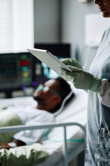 Vertical shot of female doctor checking her patient in hospital ward making notes in online medical...