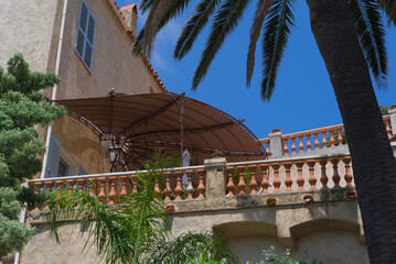 A large sunshade covers a terracotta balcony with a balustrade, with palm fronds and a clear blue...