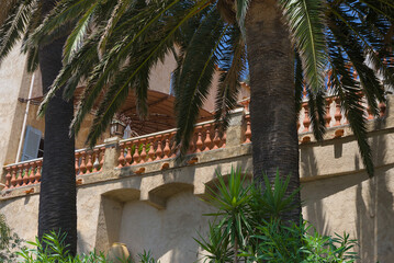 A large sunshade covers a terracotta balcony with a balustrade, with palm fronds and a clear blue...