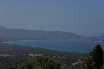 An aerial view shows a calm turquoise bay nestled between lush green hills and a distant mountain...