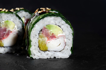 Delicious uramaki sushi rolls with tuna, avocado, surimi and cucumber filling covered with sesame seeds on black background