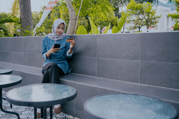 Beautiful young asian muslim female in hijab holding his smart phone and credit card, banks online or makes shopping while sitting against at outdoor cafe. Online payment concept.