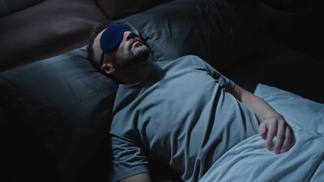 Medium shot of young Caucasian man wearing black mask lying in bed at night, falling asleep, then turning on back and adjusting eye cover to block light from window