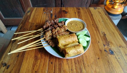 Savory satay skewers and crispy spring rolls with peanut sauce and cucumber slices on a rustic wooden table.