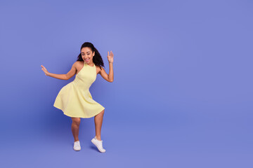 Cheerful young woman dancing joyfully in a vibrant yellow dress against a solid pastel purple background in a studio