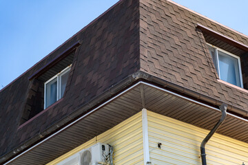 Close-up of residential building's upper corner featuring dark brown shingled mansard roof, dormer window, rain gutters and light yellow siding under clear blue sky