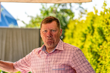 Outdoor portrait of serious-looking, middle-aged man with red hair and pink plaid shirt, displaying stern or grumpy expression against bright, blurry green and yellow natural background