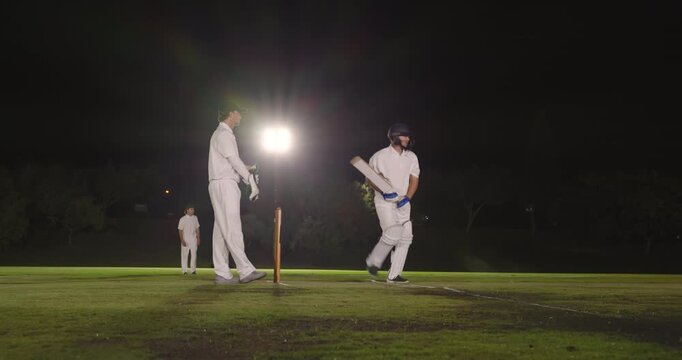 Walking onto floodlit pitch male cricketers adjusting gloves bowler starting run-up for first ball