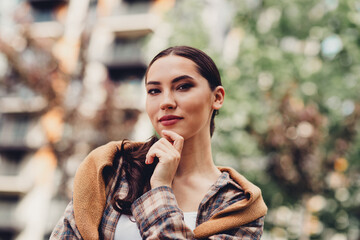Portrait of a young woman in a casual outfit standing outdoors in the city under natural light,...