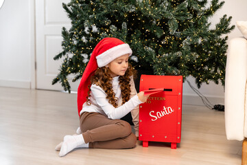 Happy little girl wearing red Santa hat sits on the floor near a Christmas tree, happily dropping a...