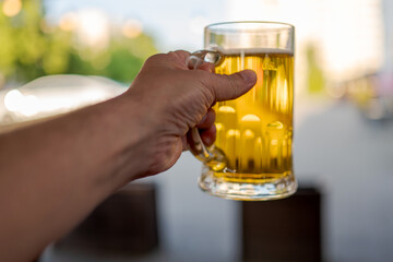 Hand holding a glass mug of beer outdoors, with blurred green background. Refreshing drink in...