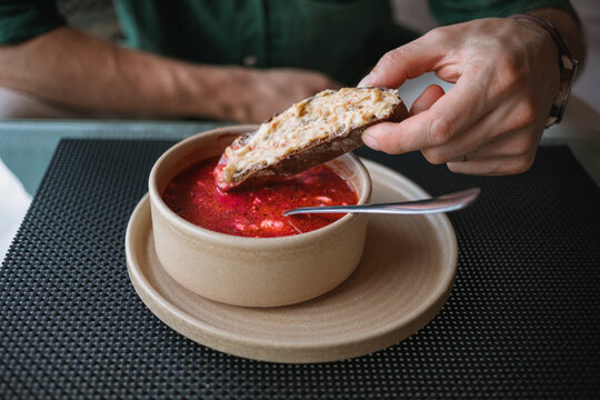 Close-up of a man sitting at a table dipping a slice of bread with garlic bacon dip into bowl of traditional borscht soup