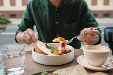 Close-up of a man sitting at a table eating shakshouka with eggs, sliced bread, a glass of water and latte coffee