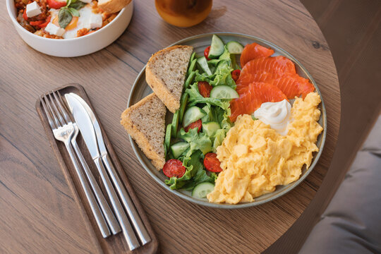 Overhead view of scrambled eggs, smoked salmon, mixed salad, multigrain bread and cream cheese on a table with a bowl of shakshouka and iced bumble coffee