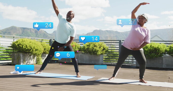 Stretching senior pair in sportswear on rooftop terrace with yoga mats and floating reaction icons