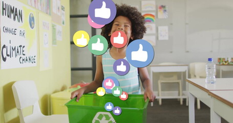 Holding rim of green recycling bin, boy standing in school classroom, with digital thumbs-up icons