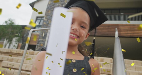 Smiling child wearing mortarboard cap and dress holding diploma on stone steps, with gold confetti
