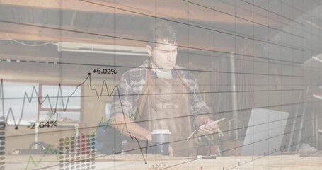 Craftsman examining tablet and viewing financial charts in carpentry workshop, with laptop, coffee