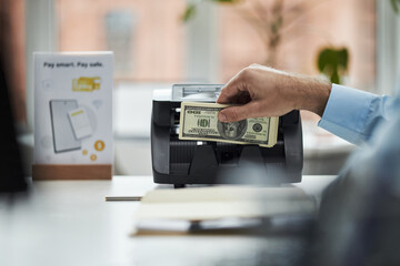 Male employee inserting US dollar bills into money counting machine in bank office, hand visible holding cash while processing transaction, blurred background with payment sign