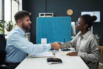 Caucasian male financial advisor shaking hands with young Black female customer across desk in bank...