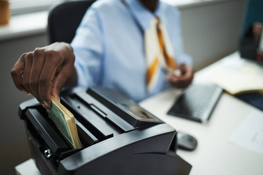 Hand of bank worker processing stack of cash using bill counting machine in office, hand inserting banknotes while sitting at desk with computer keyboard and documents visible