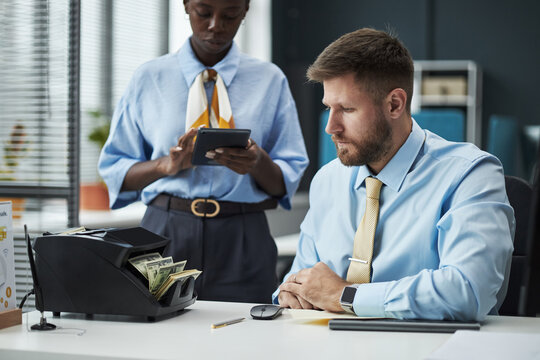Caucasian man sitting at desk operating money counting machine while Black young adult woman standing nearby using digital tablet in modern bank office environment - Powered by Adobe