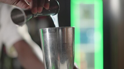 Bartender pouring liquid from jigger into cocktail shaker, focused on mixing ingredients in modern bar with green neon lighting, showcasing bartending skills and precision