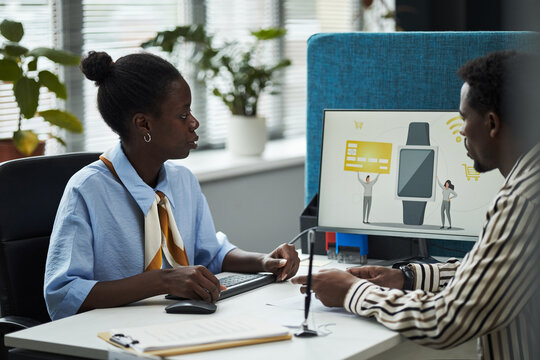 Young Black female bank worker sitting at desk discussing digital banking services with middle aged man in modern financial office, computer monitor displaying online payment interface