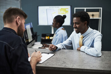 Black middle aged man wearing business attire, assisting Caucasian young man filling out paperwork...