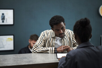 Young Black man leaning on counter, listening to middle aged Black woman explaining information in bank office setting, with another Caucasian male customer waiting in queue in background