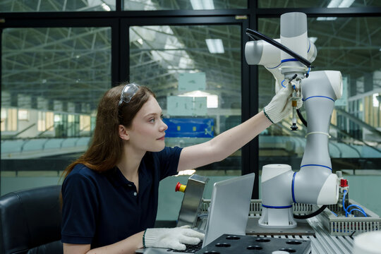 female robotics trainee adjusting robotic arm in smart factory using laptop and manual control system during training session focusing on workflow accuracy and hands-on skill learning