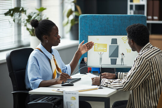 Young Black female consultant explaining digital banking services to male customer in bank office, both sitting at desk with computer monitor displaying financial technology graphics