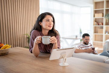 Woman drinking coffee and using tablet at home while man relaxes on sofa