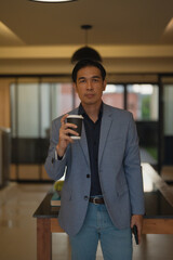 Asian businessman dressed in smart casual attire, holding a takeaway coffee and smartphone,...