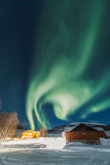 Northern lights light up the entire sky under small wooden cottages with fire inside. Cozy and...