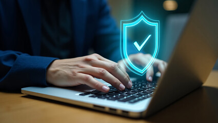 Hands type on a laptop, with a glowing blue shield icon (checkmark) floating above, representing cybersecurity or data protection.