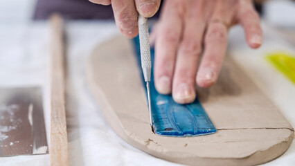 A close-up shot of a middle-aged woman artist shaping clay with sculpting tools at a table in an art studio, with an environment adorned with artistic paintings in the background
