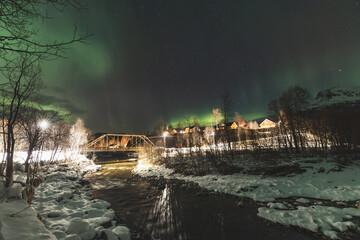 Aurora Borealis glows above river and village in Troms region, painting the starry night with green...