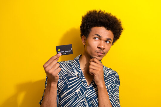 Young man holding a credit card while thinking against a yellow background, wearing patterned shirt with a thoughtful look